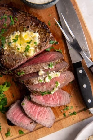 A top down shot of a partially sliced London Broil topped with melting blue cheese butter on a wood board with a knife and serving fork.