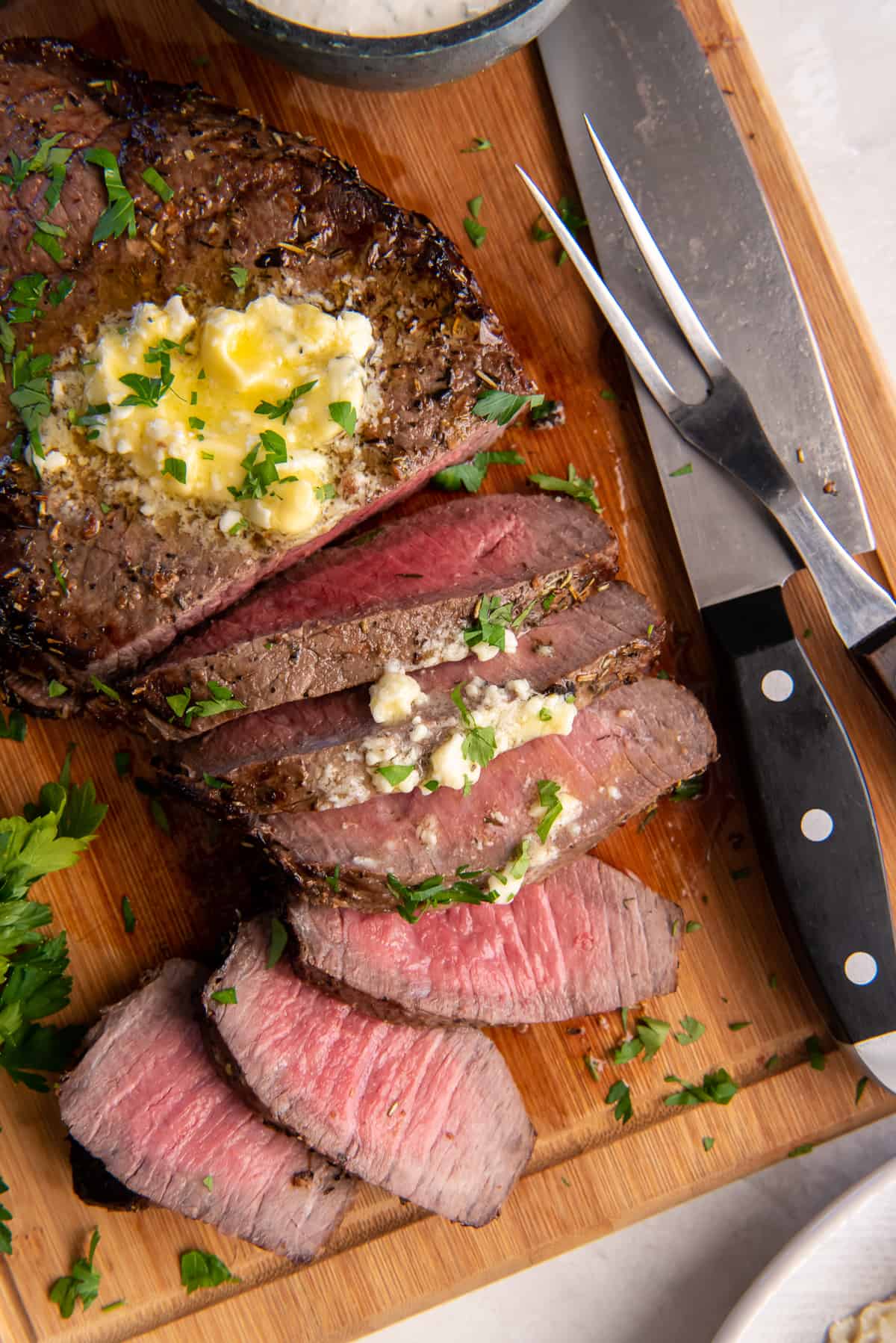 A top down shot of a partially sliced London Broil topped with melting blue cheese butter on a wood board with a knife and serving fork.