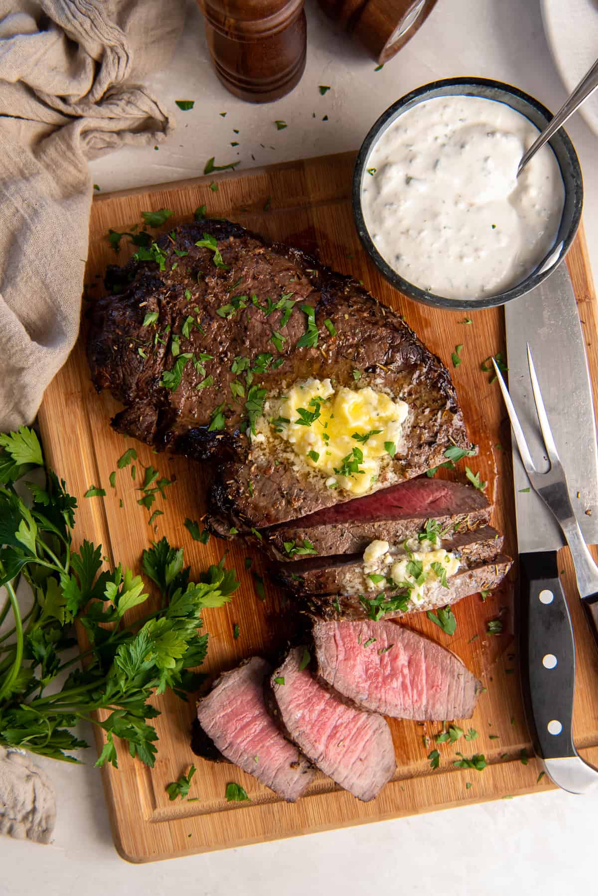 A top down shot of a partially sliced London Broil topped with melting blue cheese butter on a wood board with a small bowl of a creamy sauce.