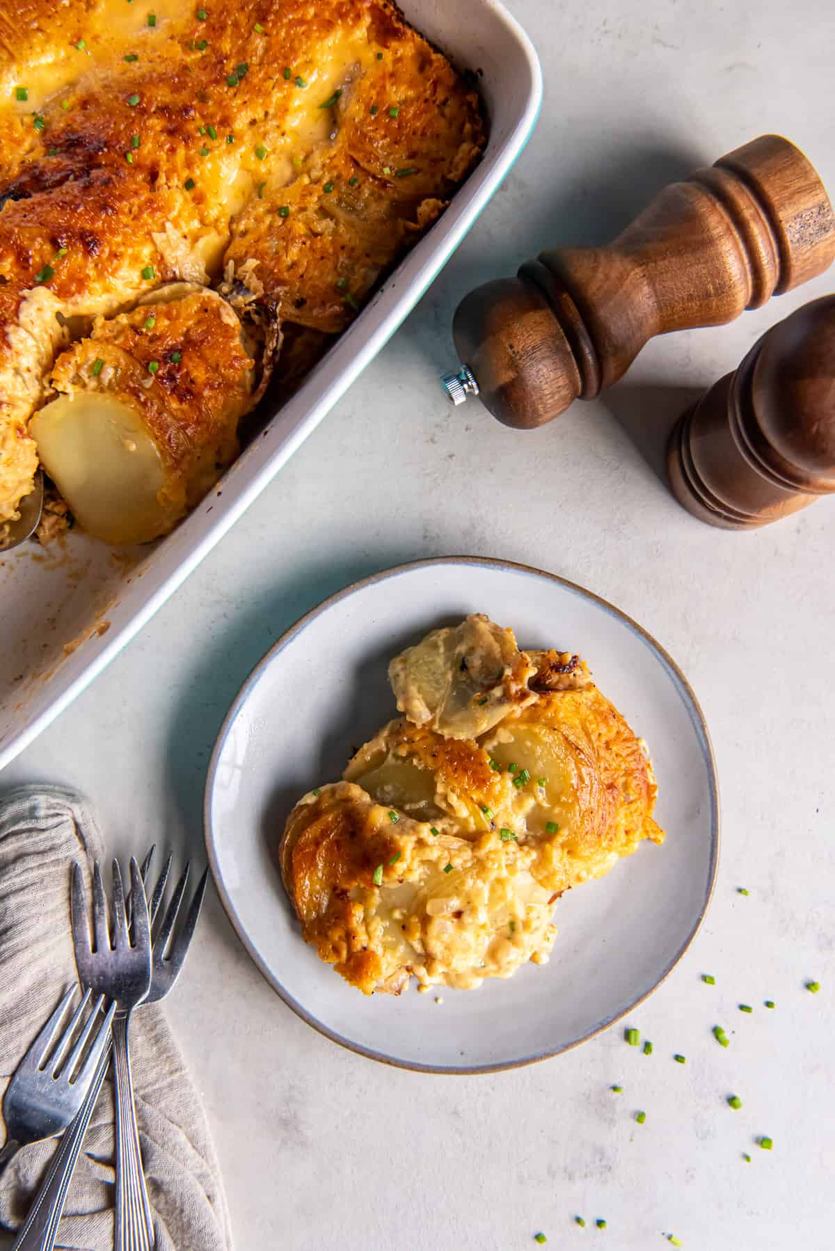 A top down shot of a serving of cheesy au gratin potatoes on a white plate next to salt and pepper shakers and a baking dish full of the potatoes.