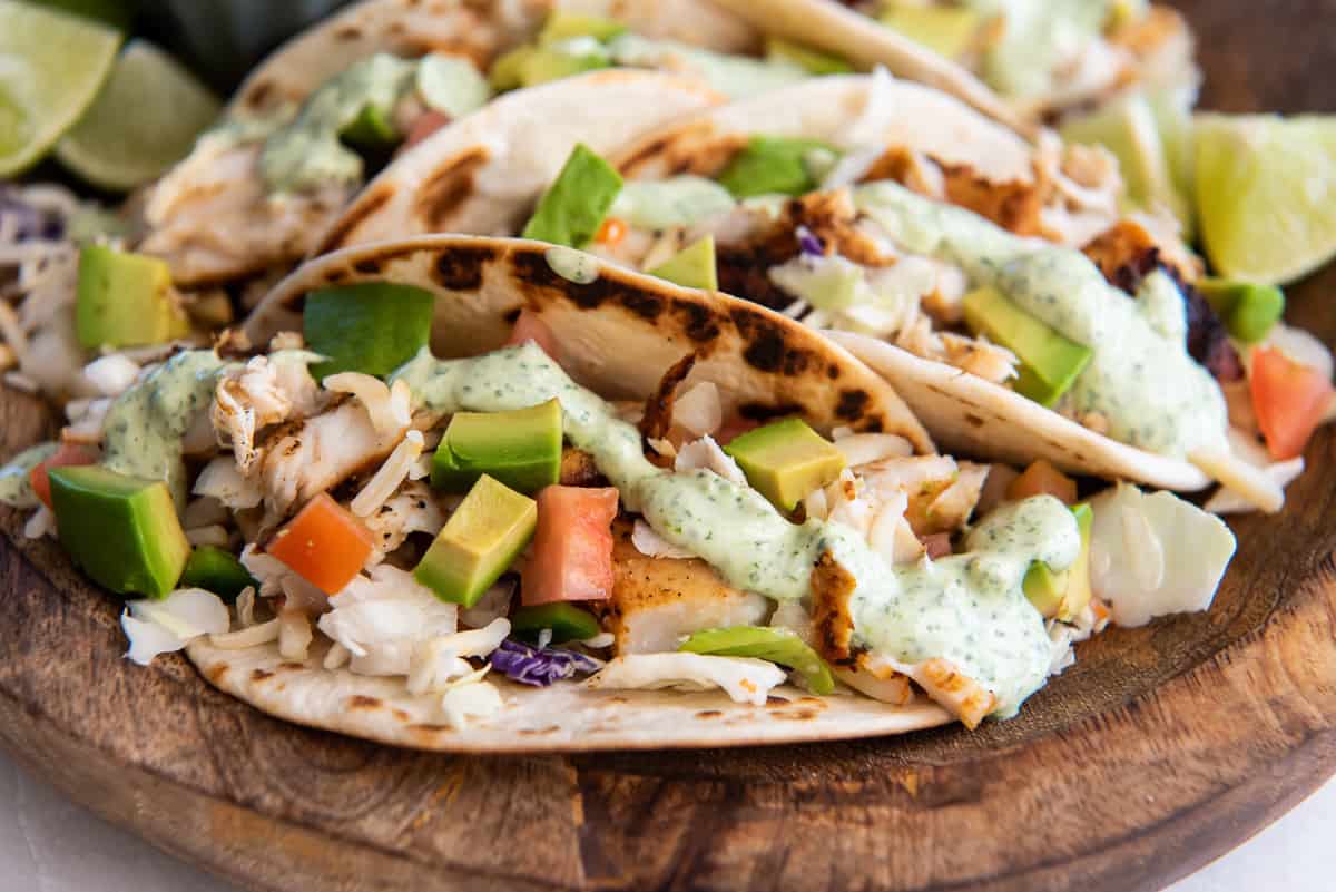 An eye level shot of a blackened fish taco on a flour tortilla with tomatoes, avocado and cilantro lime sauce on a wood platter.