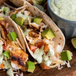 Blackened fish tacos with cabbage and avocado on a wood platter with a small bowl of cilantro lime sauce.