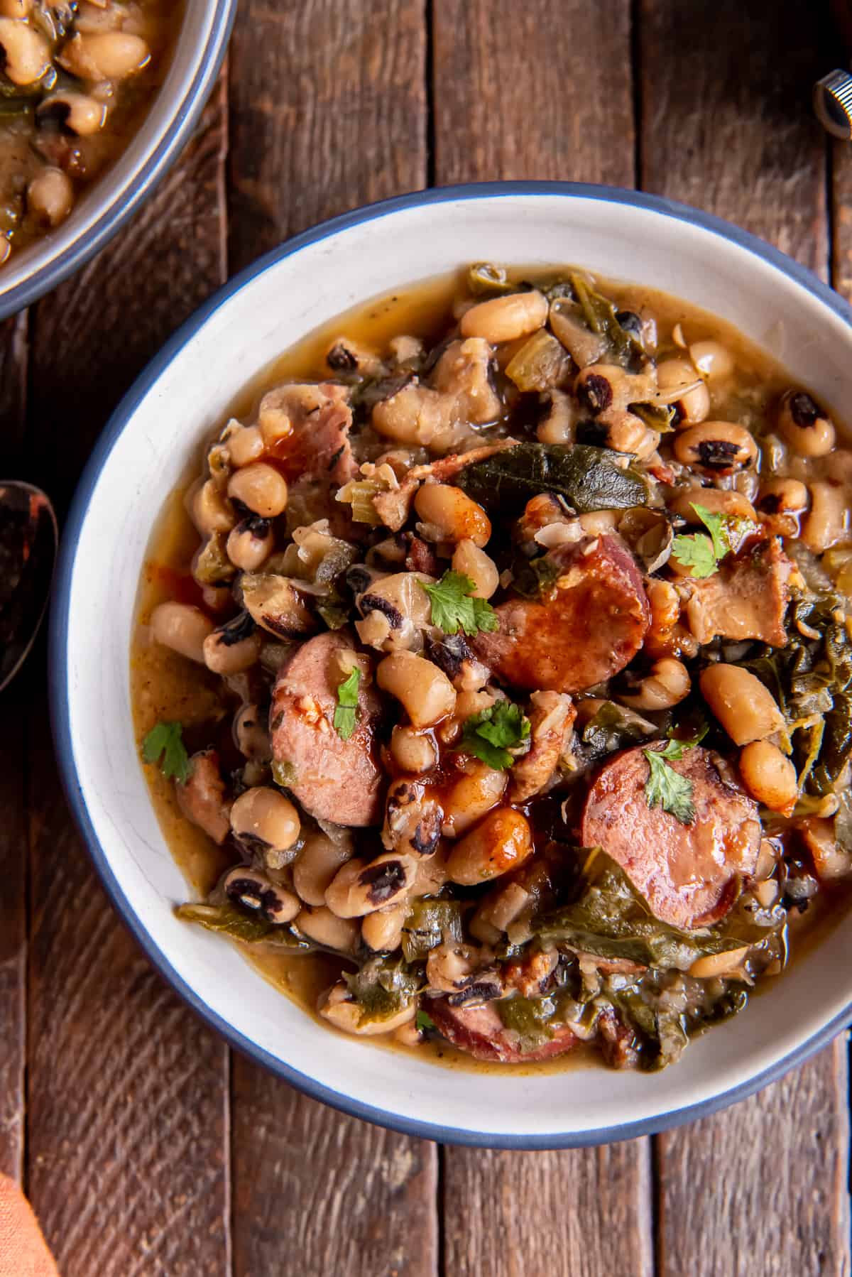 A top down shot of a bowl filled with black eyed peas and sausage with collard greens on a dark wood board.