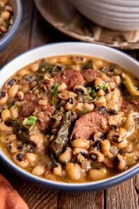 Two bowls of black eyed peas with sausage and collards on a wood board.