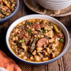 Two bowls of black eyed peas with sausage and cabbage on a wooden board.