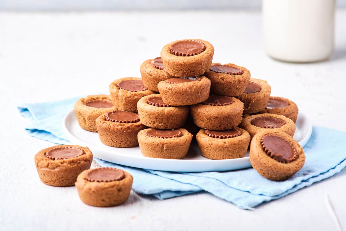 Cookies with peanut butter cups in the center piled on a white plate on a blue cloth with two on the counter in front of it.