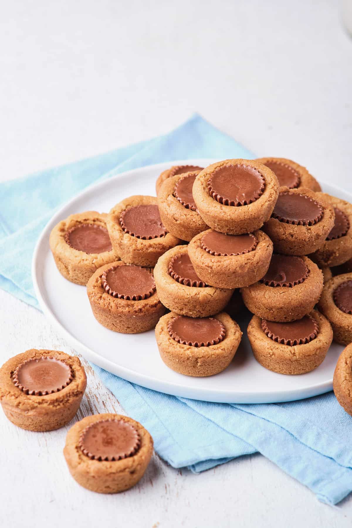 Cookies with peanut butter cups in the center piled on a white plate on a blue cloth with two on the counter in front of it.