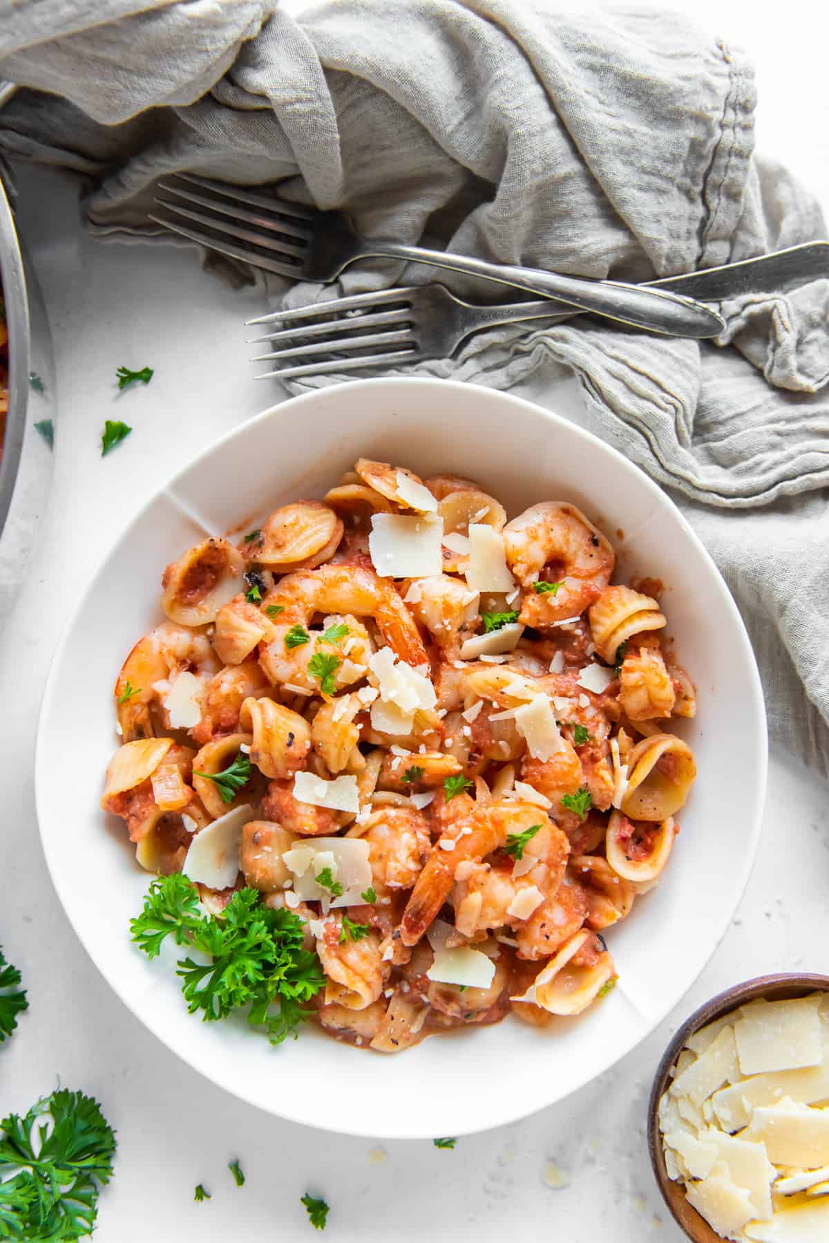 A top down shot of a white bowl filled with shrimp pasta in vodka sauce garnished with Parmesan and parsley next to a grey cloth and forks.