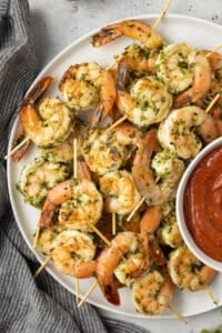 A top down shot of seared shrimp skewers on a white platter with cocktail sauce.