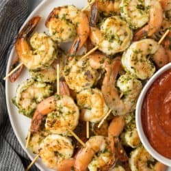 A top down shot of seared shrimp skewers on a white platter with cocktail sauce.