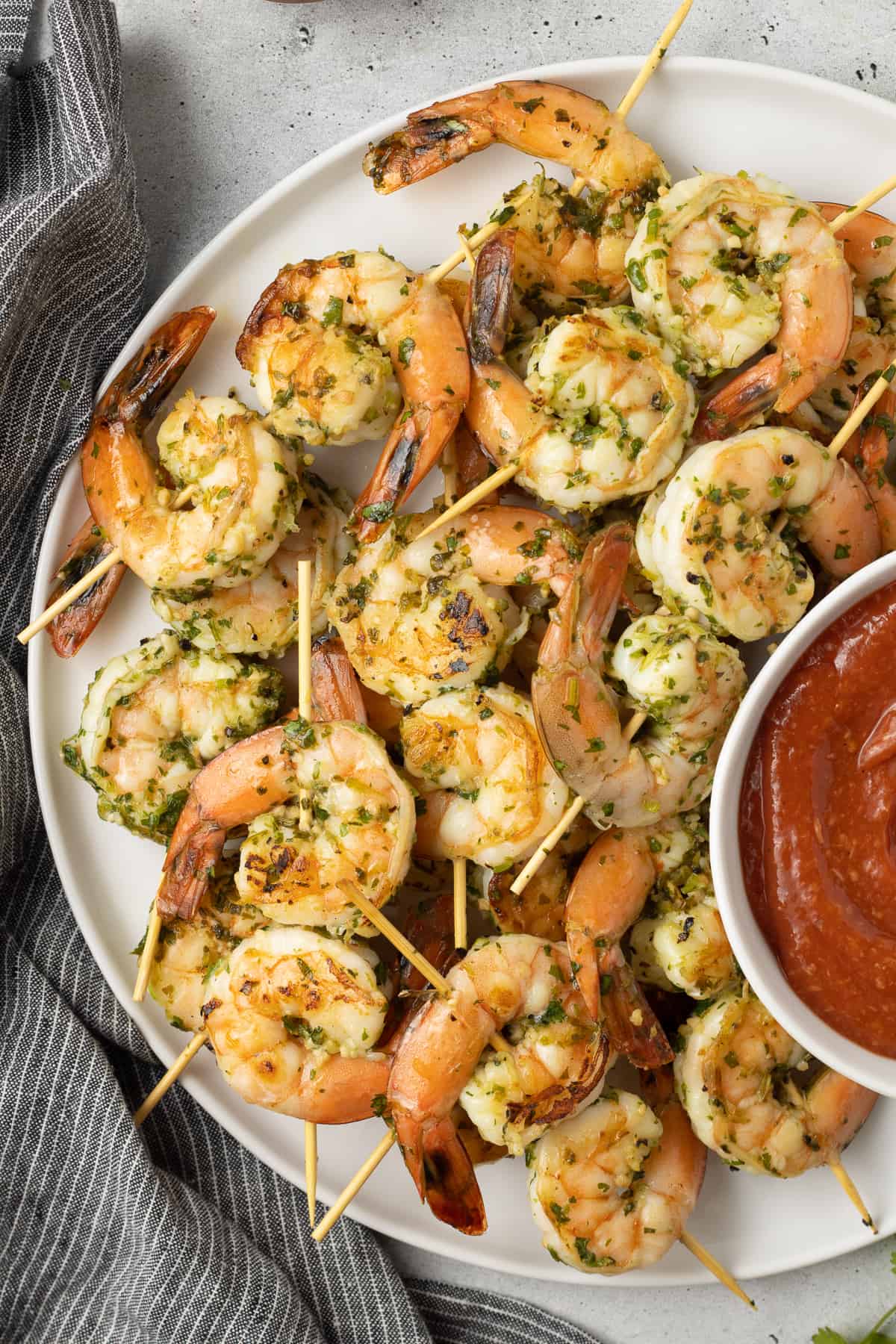 A top down shot of seared shrimp skewers on a white platter with cocktail sauce.