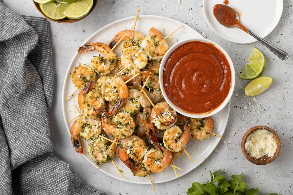 Shrimp skewers on a platter with cocktail sauce next to a small bowl of cotija cheese and lime wedges.