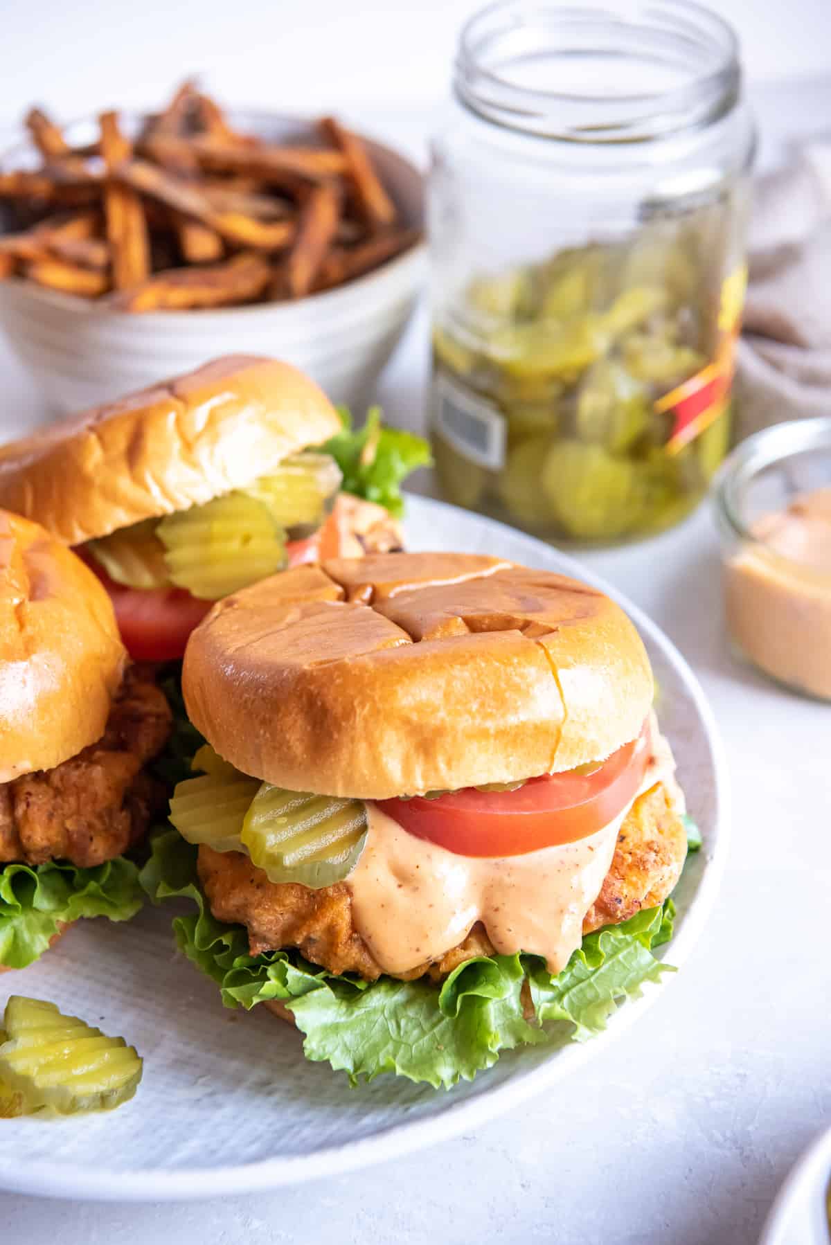 Pickle brined chicken sandwiches on a platter in front of a jar of pickles and a bowl of French fries.