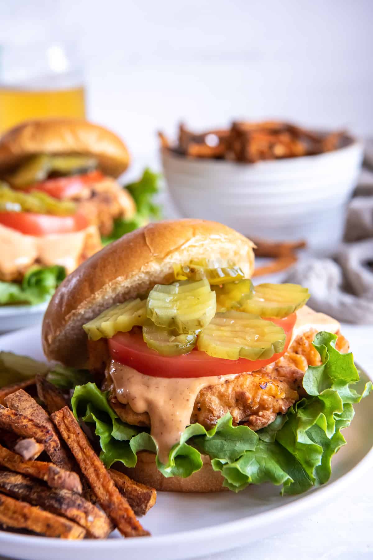 A crispy chicken sandwich topped with sauce and pickles with the top bun offset on a plate with fries.