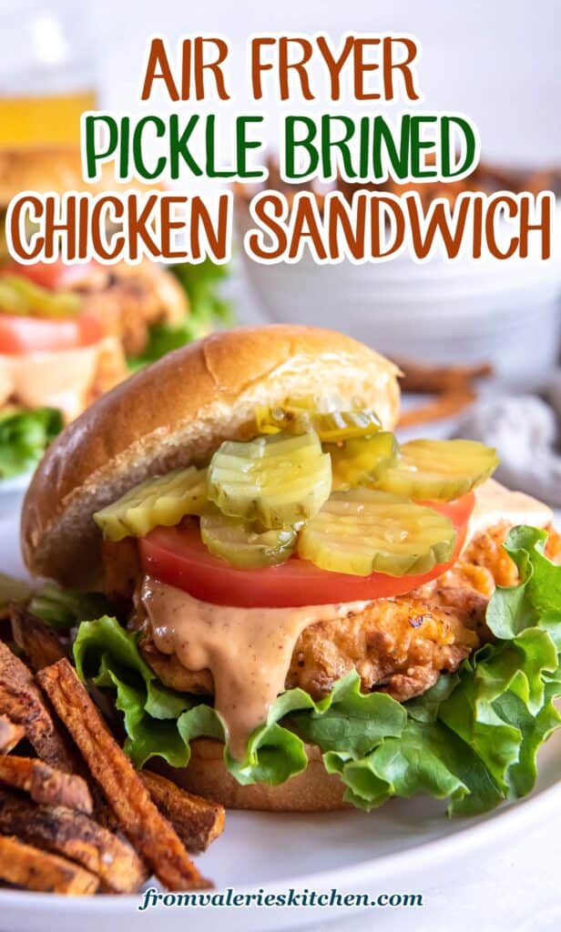 A crispy chicken sandwich topped with sauce and pickles with the top bun offset on a plate with fries with text.