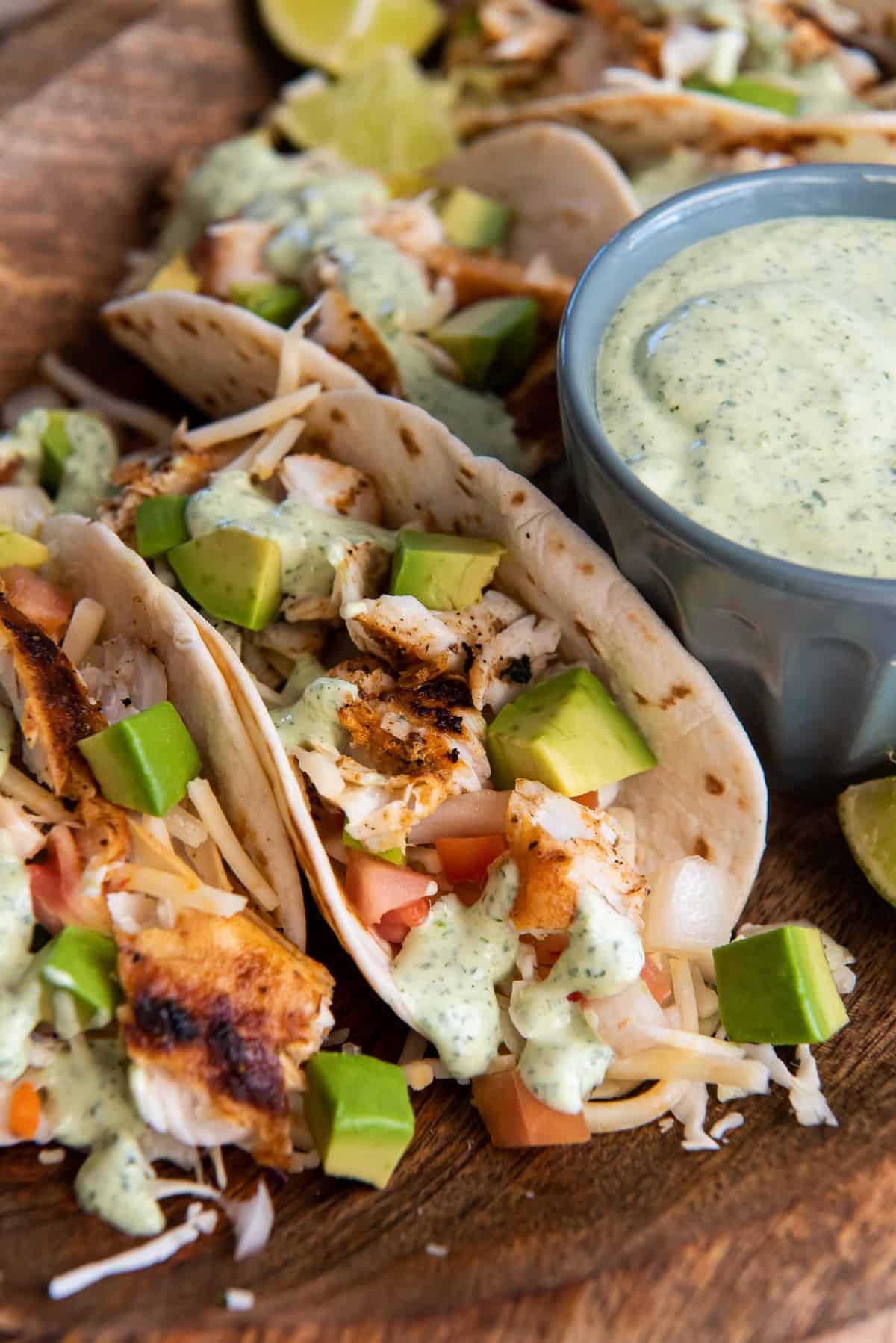 Blackened fish tacos with cabbage and avocado on a wood platter with a small bowl of cilantro lime sauce.