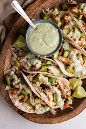 A top down shot of blackened fish tacos on a wood board with a small bowl of cilantro lime sauce.