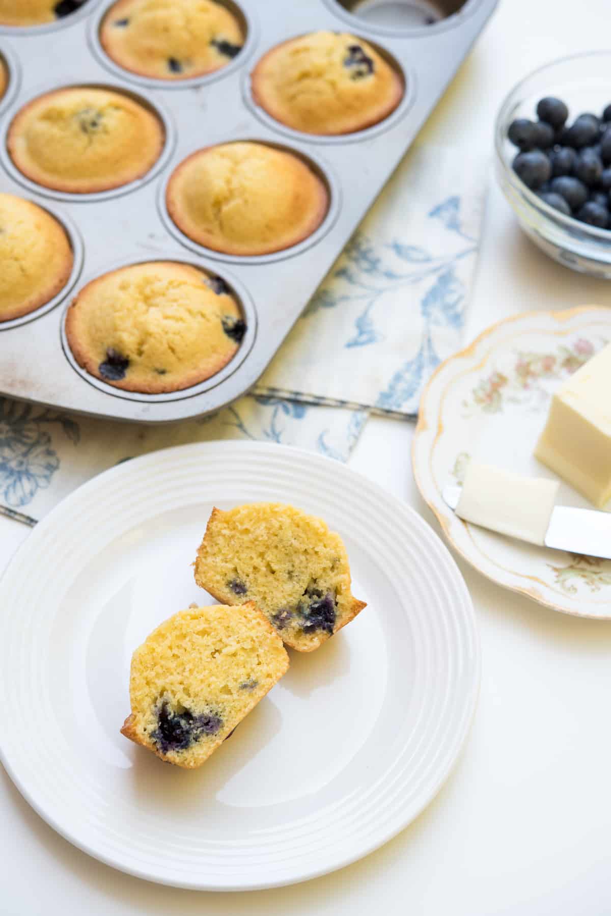 A blueberry cornmeal muffin sliced in half on a white plate next to a small plate with butter and a knife.