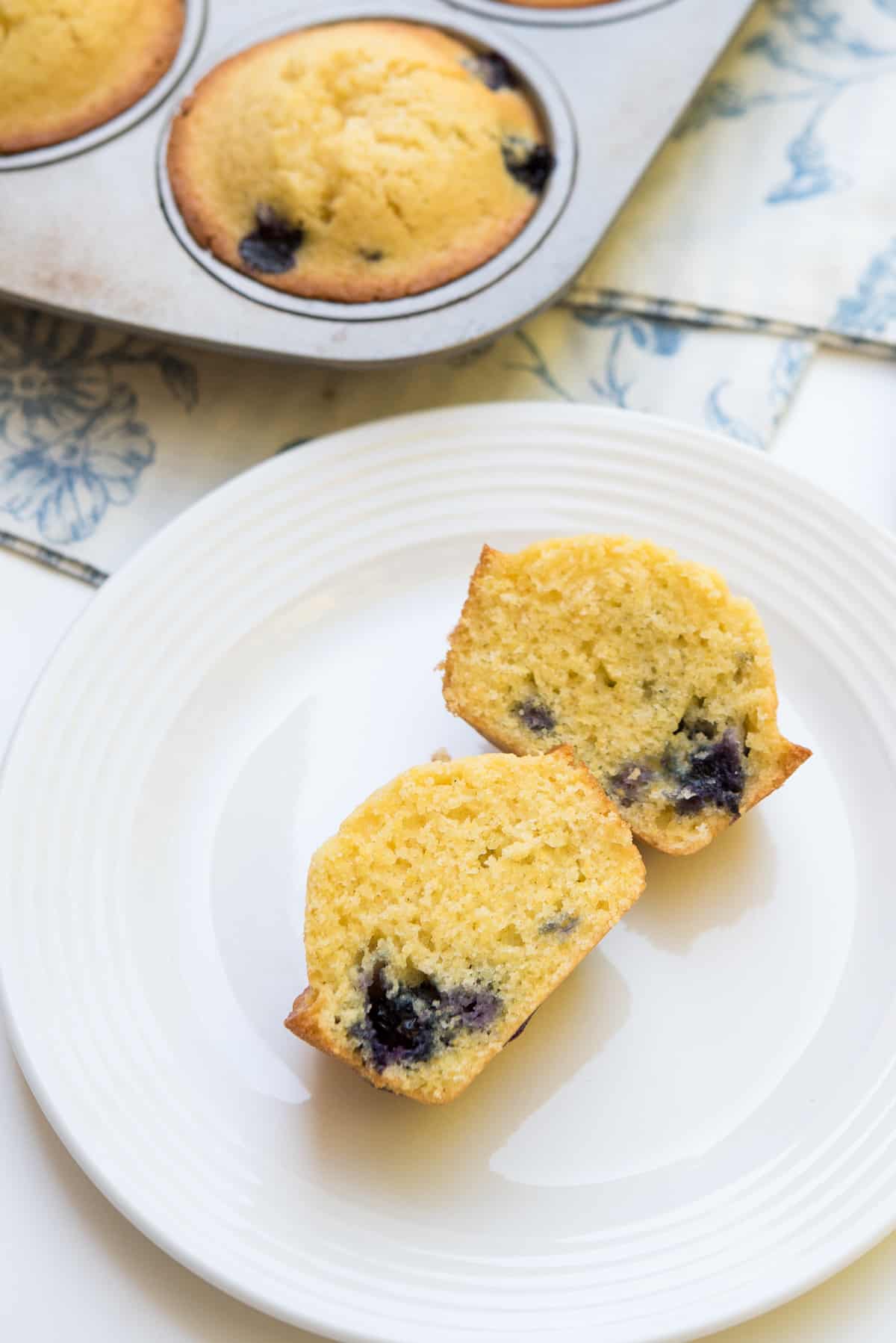 A blueberry cornmeal muffin sliced in half on a white plate next to a muffin tin.