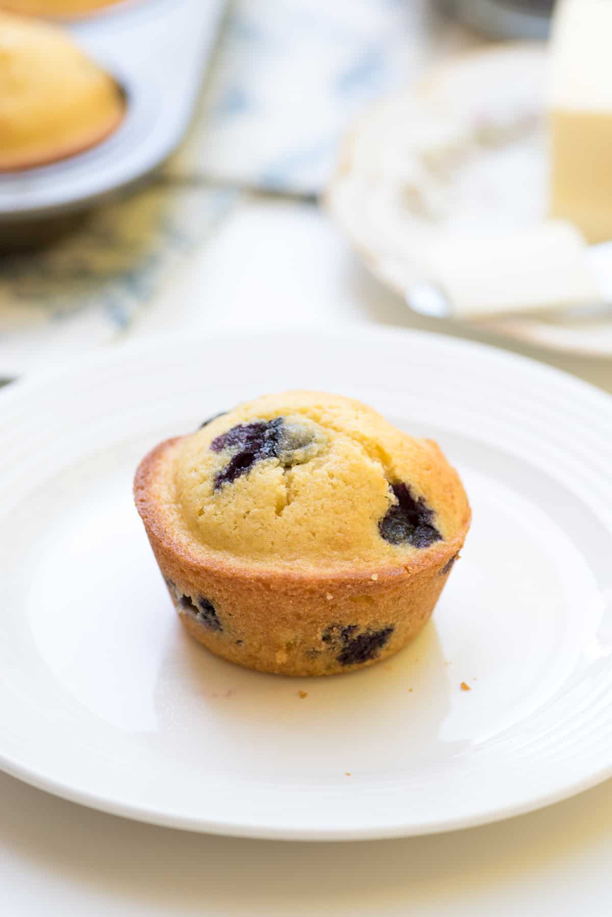 A cornmeal muffin with blueberries on a white plate with a muffin tin and plate with butter in the background.