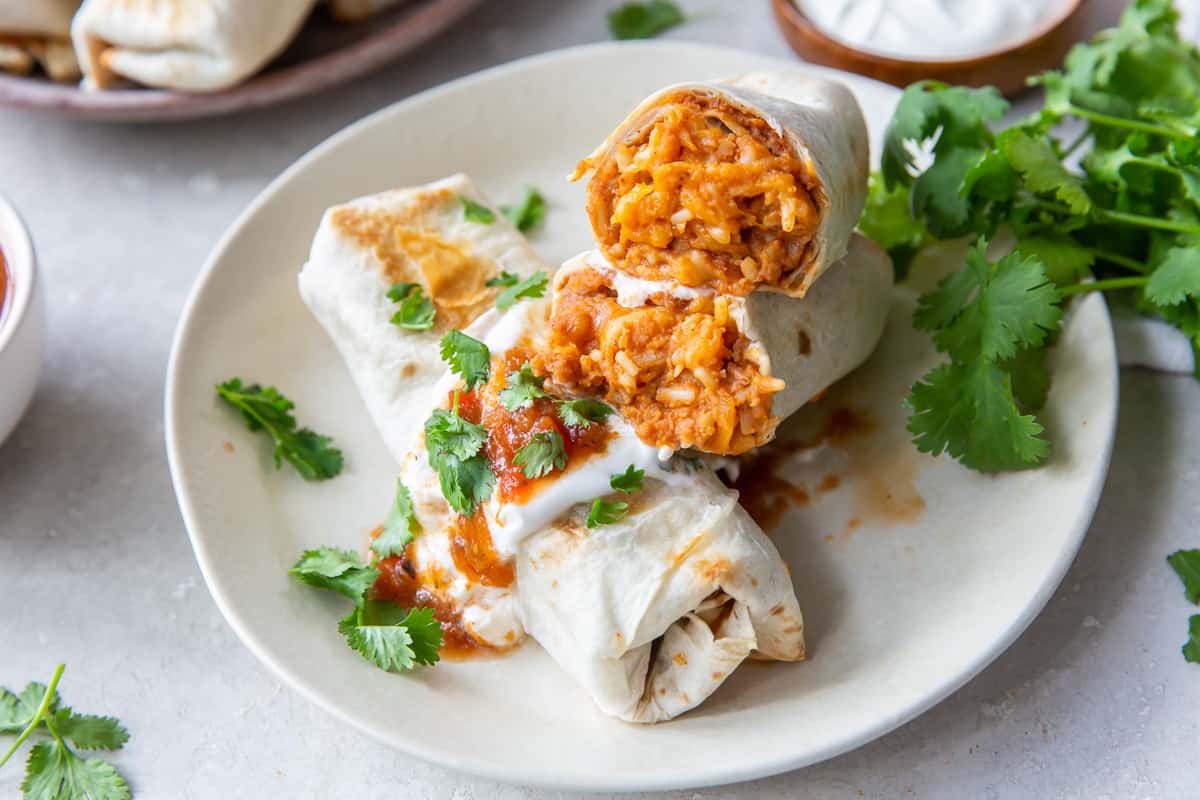 A burrito is sliced in half to reveal beans, rice, and cheese and stacked on top of another burrito with sour cream and salsa.