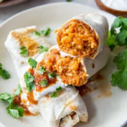 A burrito is sliced in half to reveal beans, rice, and cheese and stacked on top of another burrito with sour cream and salsa.