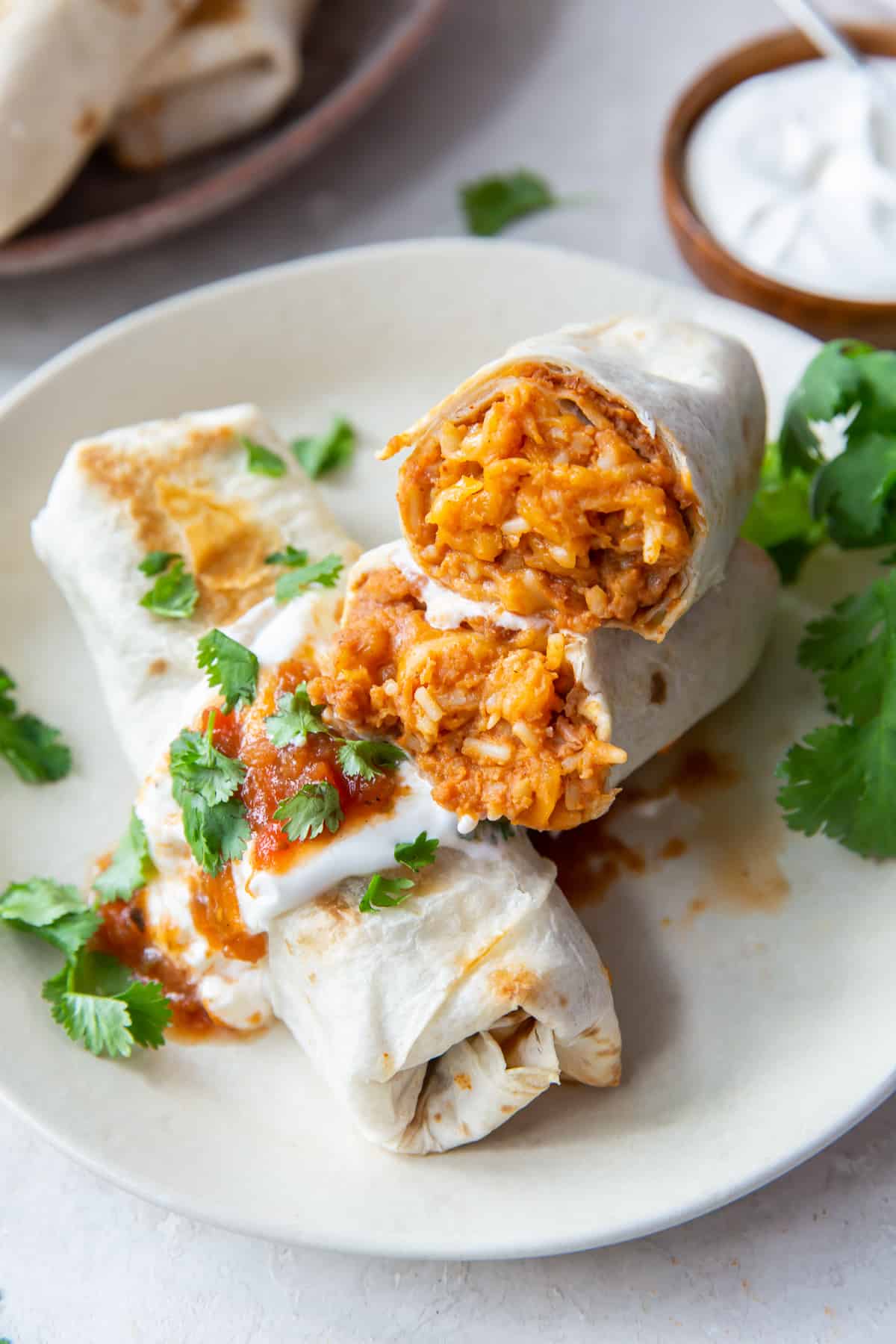 A burrito is sliced in half to reveal beans, rice, and cheese and stacked on top of another burrito with sour cream and salsa.