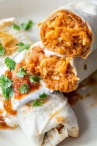 A burrito is sliced in half to reveal beans, rice, and cheese and stacked on top of another burrito with sour cream and salsa.