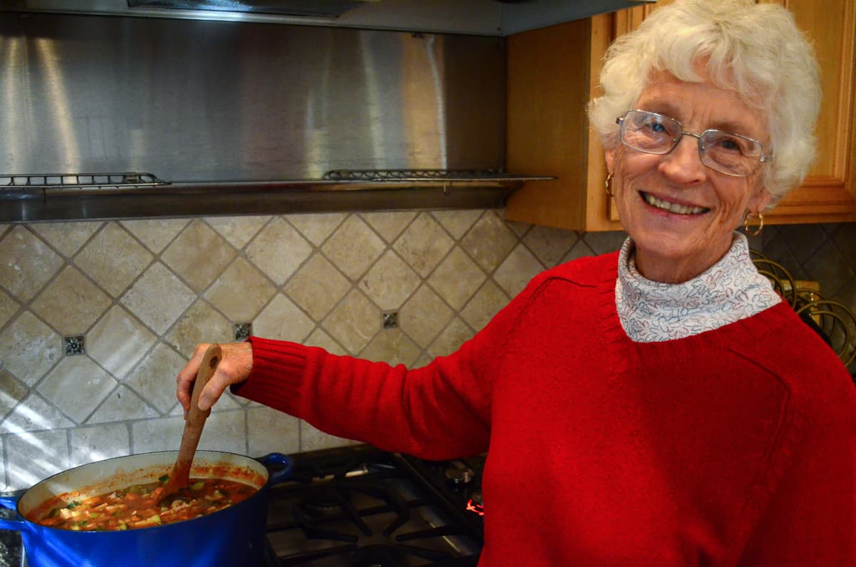 An smiling elderly woman with white hair stirring a pot of soup on the stove.