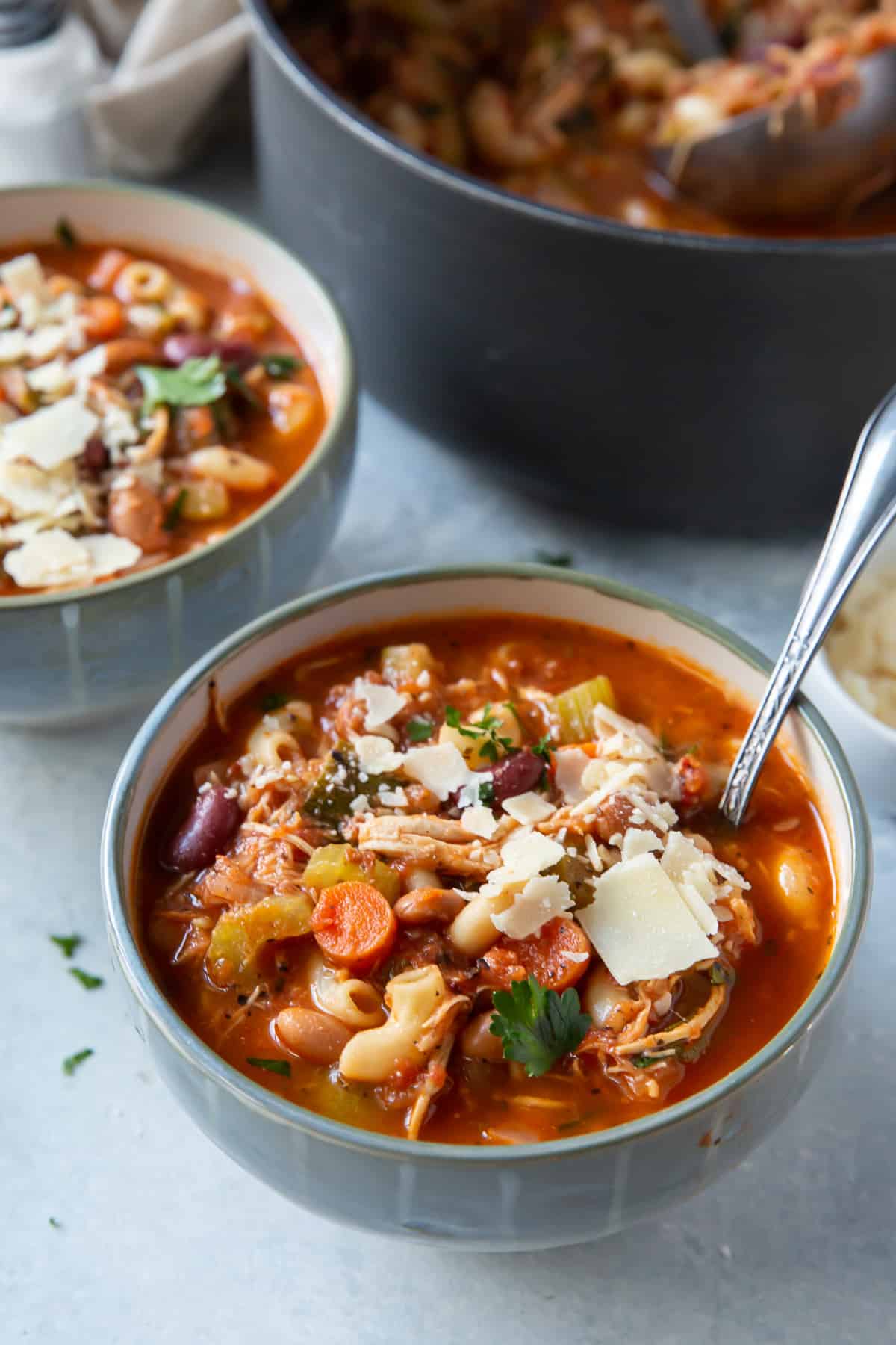 A spoon resting in a bowl of minestrone soup with shredded chicken and pasta.