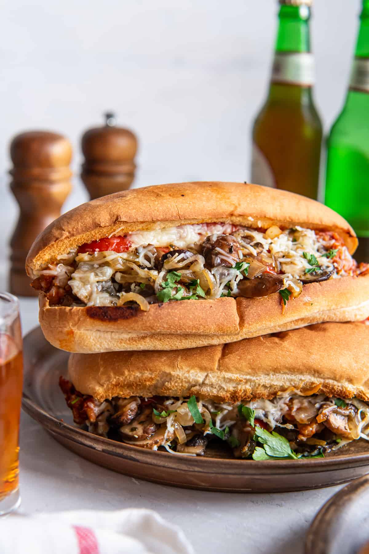 A stack of two Italian Sausage Sandwiches on toasted hoagie rolls on a platter in front of beer bottles and salt and pepper shakers.