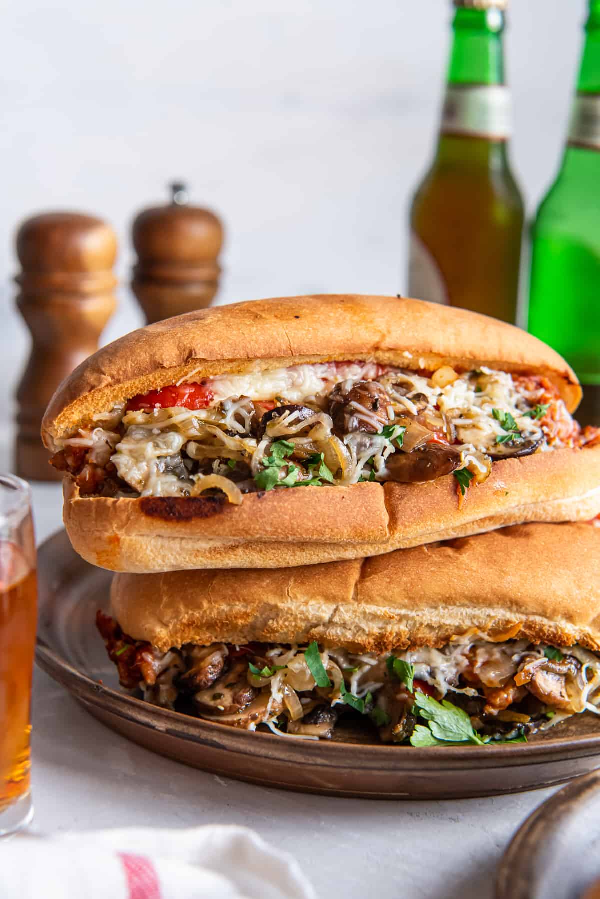 Ein Stapel von zwei italienischen Wurstsandwiches auf gerösteten Hoagie-Brötchen auf einer Platte vor Bierflaschen und Salz- und Pfefferstreuern.