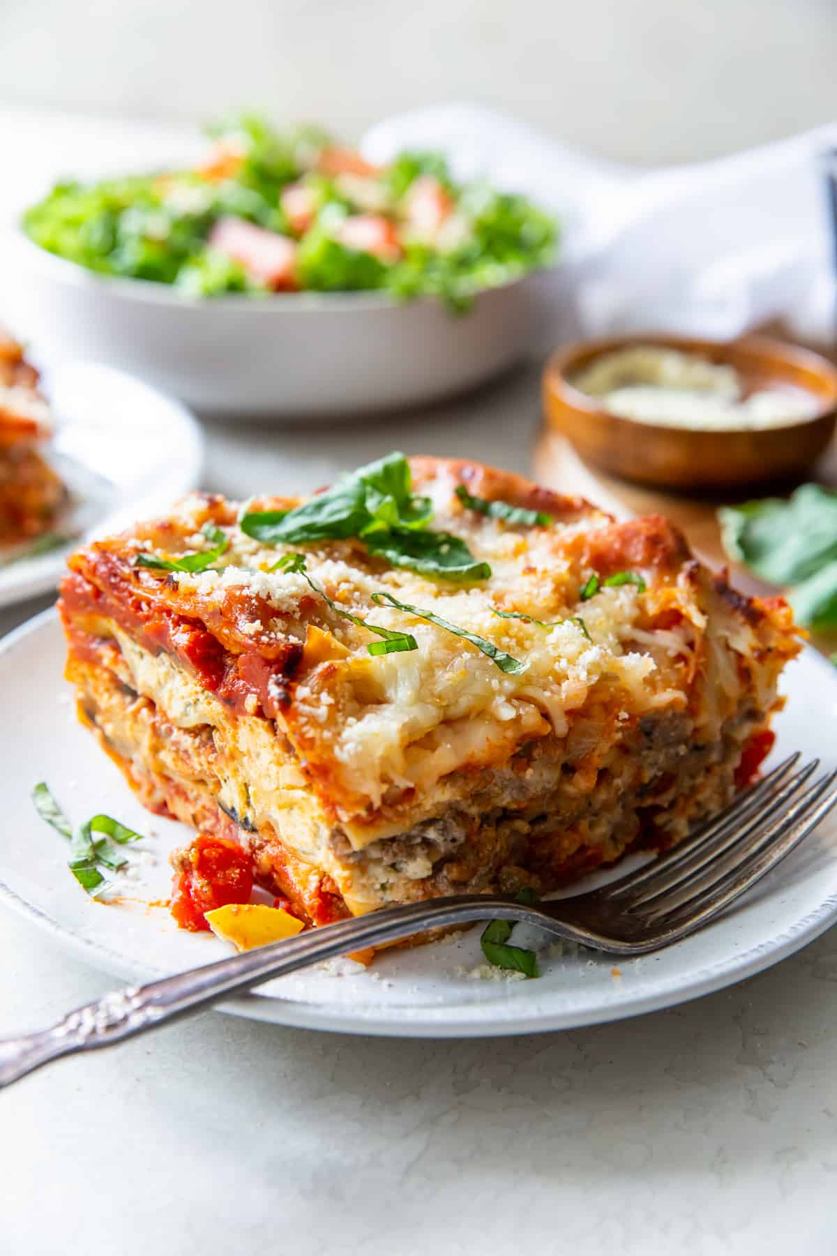 A piece of lasagna topped with fresh basil on a white plate with a green salad in the background.