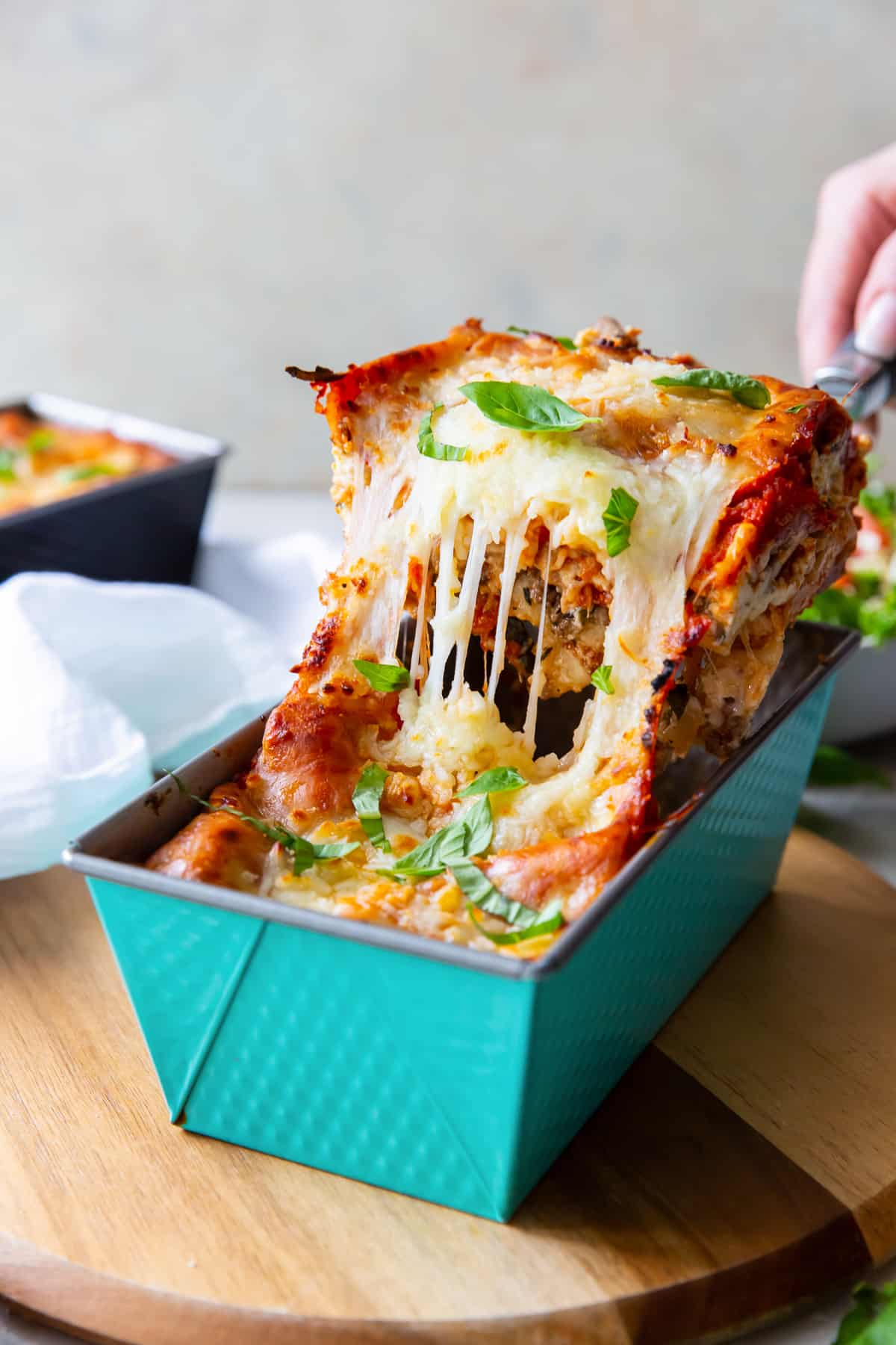 A hand holding a spatula lifting a piece of lasagna from a blue metal loaf pan.