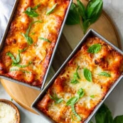 A top down shot of lasagna in two loaf pans with fresh basil and a small bowl of Parmesan around them.