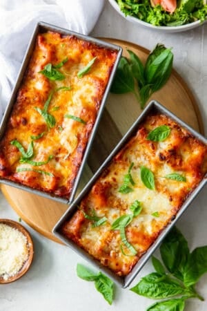 A top down shot of lasagna in two loaf pans with fresh basil and a small bowl of Parmesan around them.