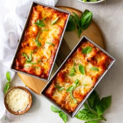 A top down shot of lasagna in two loaf pans with fresh basil and a small bowl of Parmesan around them.