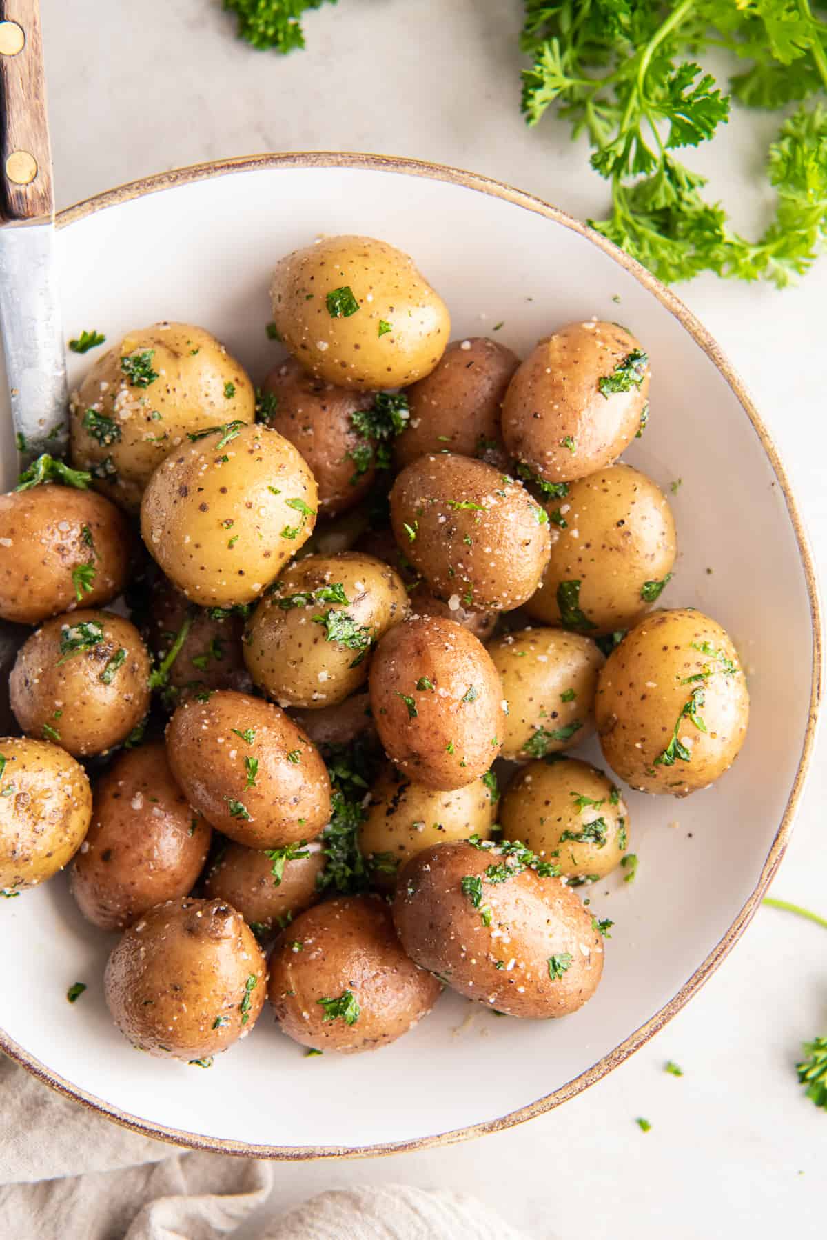 A top down shot of parsley potatoes in a white bowl with fresh parsley around it.