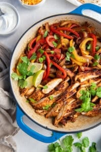 A top down shot of chicken fajitas in a skillet with cilantro and lime wedges next to a small bowl of sour cream and cheese.