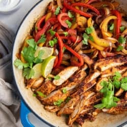 A top down shot of chicken fajitas in a skillet with cilantro and lime wedges next to a small bowl of sour cream and cheese.