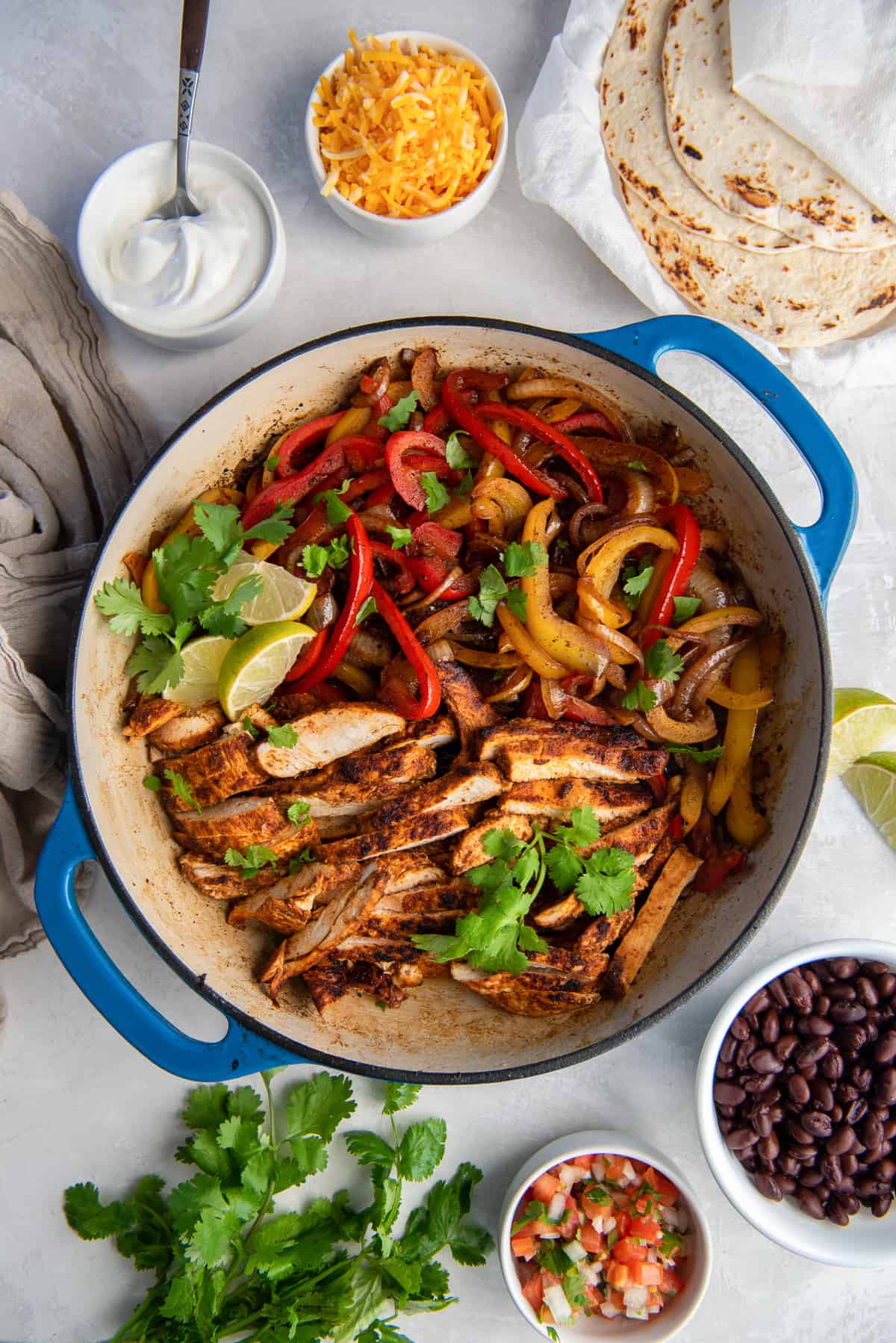 A top down shot of chicken fajitas in a skillet surrounded by sour cream, cheese, cilantro, salsa, tortillas, and black beans.