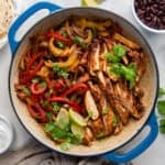 A top down shot of chicken fajitas in a skillet surrounded by sour cream, cheese, cilantro, salsa, tortillas, and black beans.