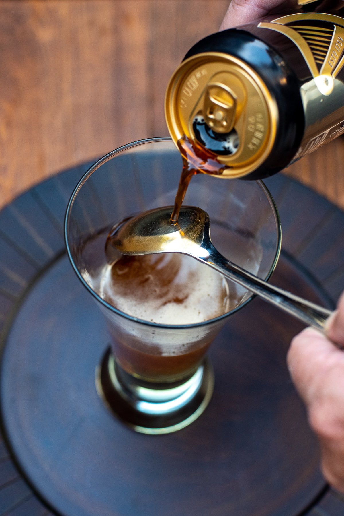 Guinness being poured from a can over a spoon and into a beer glass on top of a hard cider.