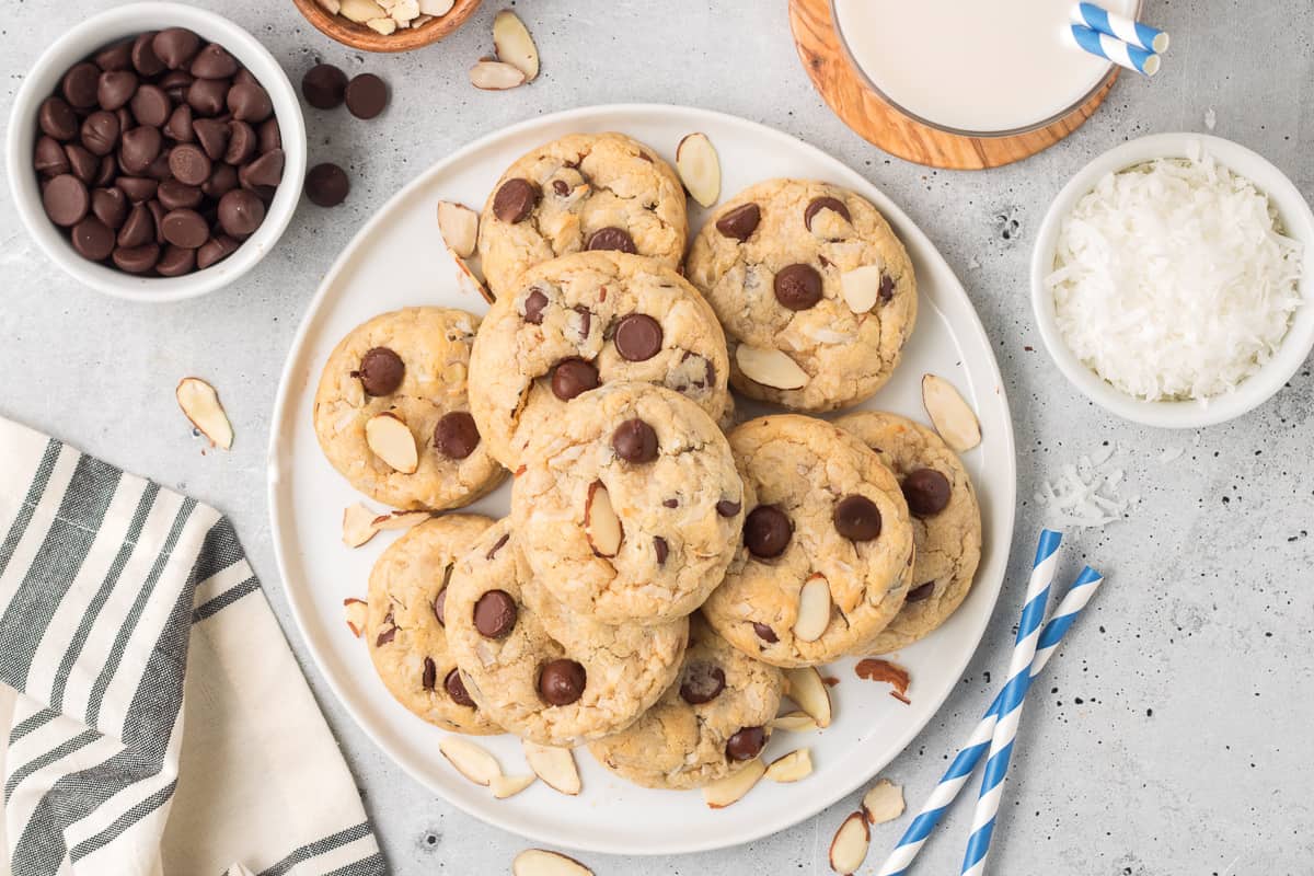 A top down shot of a white plate full of cookies with small bowls of chocolate chips, coconut and almonds around them.