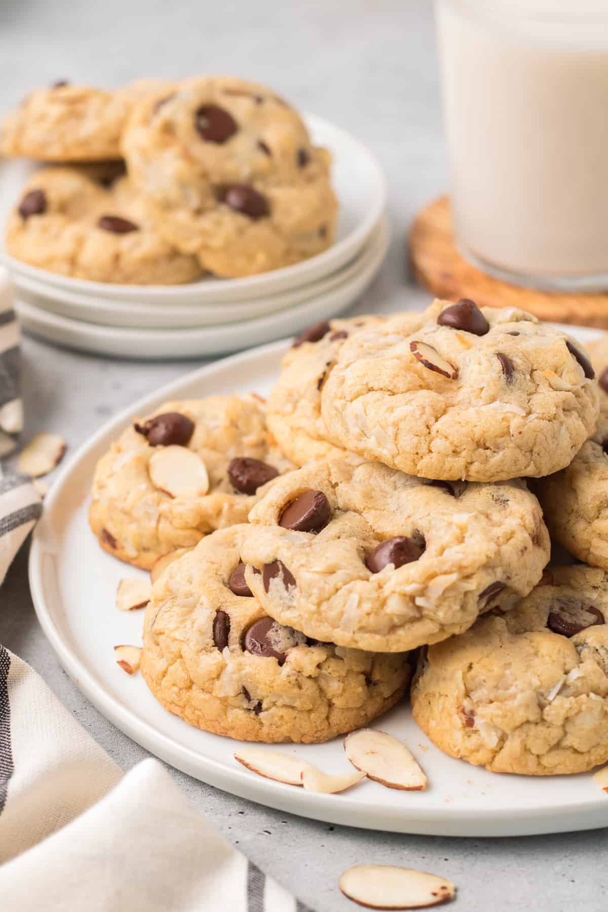 A pile of Almond Joy Cookies on a white plate with smaller plate and glass of milk in the background.