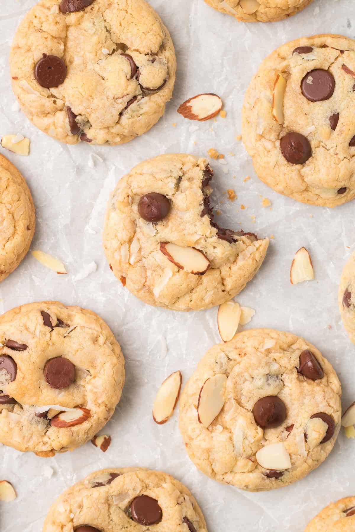 A top down shot of cookies with chocolate chips, coconut, and almonds on parchment paper. The cookie in the center is missing a bite.