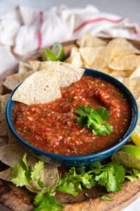 Two tortilla chips resting in a bowl of salsa on a platter with cilantro and more chips.