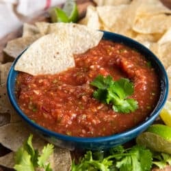 Two tortilla chips resting in a bowl of salsa on a platter with cilantro and more chips.