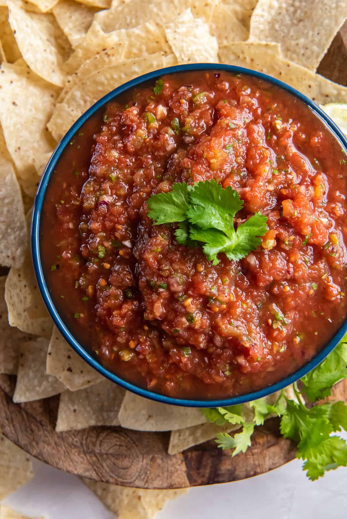 A top down shot of salsa in a bowl on a wood platter with tortilla chips.