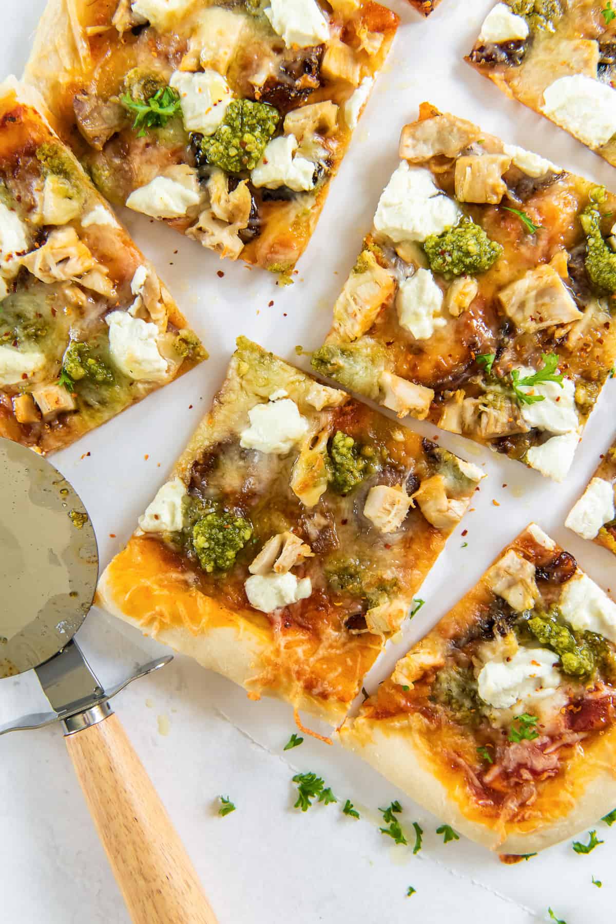 A top down shot of square pizzas of chicken pesto pizza next to a pizza cutter.